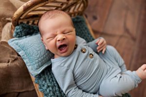 Baby in blue outfit yawning while lying on textured blue pillow.