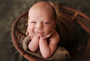 Baby posed in wooden bowl with hands under chin on dark backdrop.