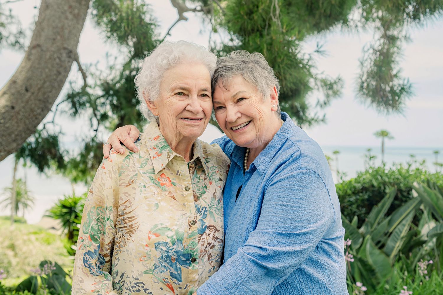 Two smiling elderly women embracing outdoors with trees and ocean in the background.