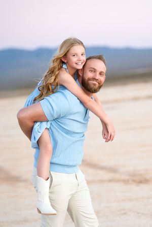 A bearded man gives a smiling blonde girl a piggyback ride on a sandy beach, both wearing light blue outfits.