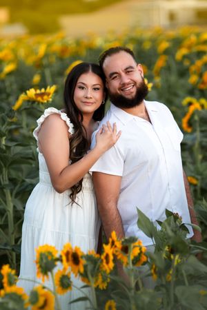 Couple poses together in field of blooming sunflowers during golden hour.