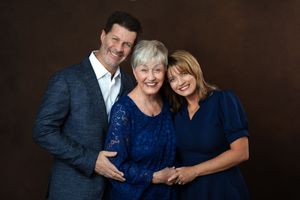Three people pose warmly together in a professional studio portrait, wearing elegant blue outfits against a brown background.