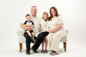 Family of four posing for a professional portrait on a white chair against a white background in matching neutral outfits.