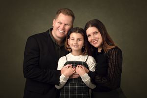 A smiling family of three poses for a studio portrait, with parents flanking their young daughter against a dark green background.