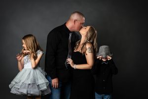 Family portrait with parents kissing, daughter in blue dress, and son hiding face, against dark gray background.