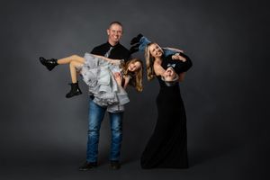 A man in jeans holds a girl in a ruffled dress while a woman in black gown poses playfully in a studio.