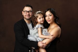 Family portrait of a man in glasses, woman in black dress, and toddler girl in blue dress with floral headband.