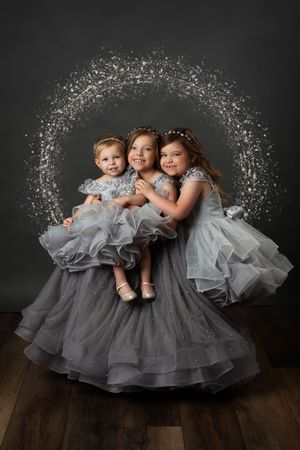 Three young sisters in matching silver tulle gowns pose together with a sparkling light halo backdrop in a studio portrait.