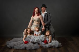 Elegant family portrait with redhead woman, man in fedora, and three girls in white dresses on studio backdrop.