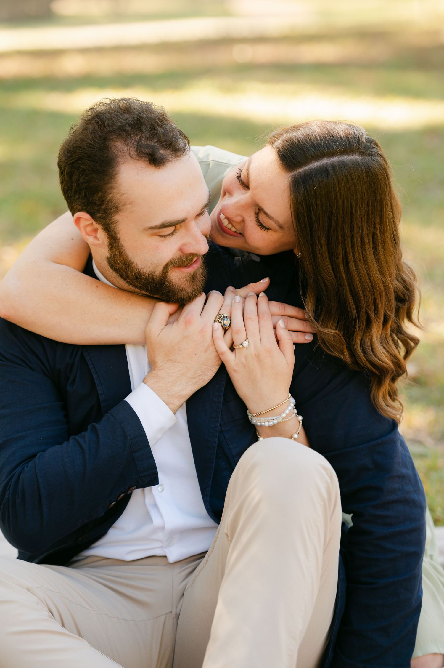 A happy couple sits on grass, embracing and showing off an engagement ring.
