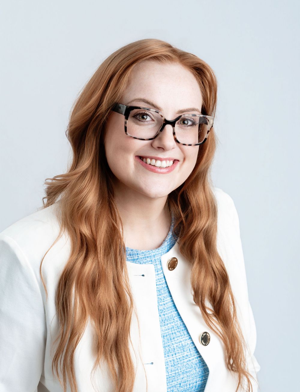 A person wearing glasses and a white cardigan smiles warmly against a light background.