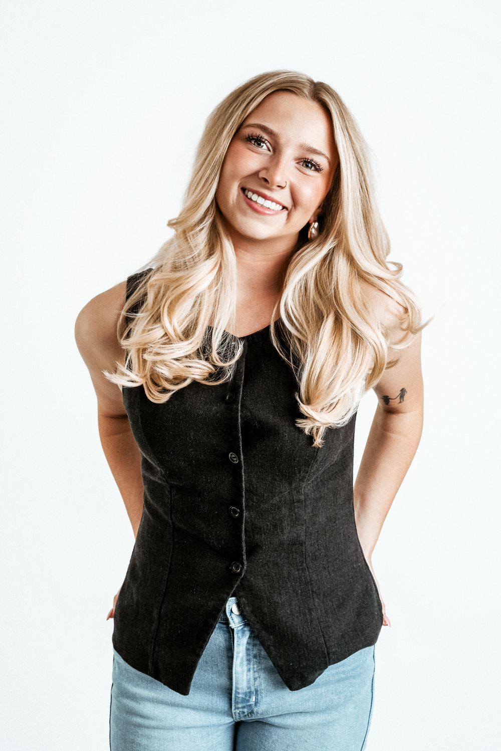 Smiling blonde woman in black vest and jeans posing confidently against a white background.