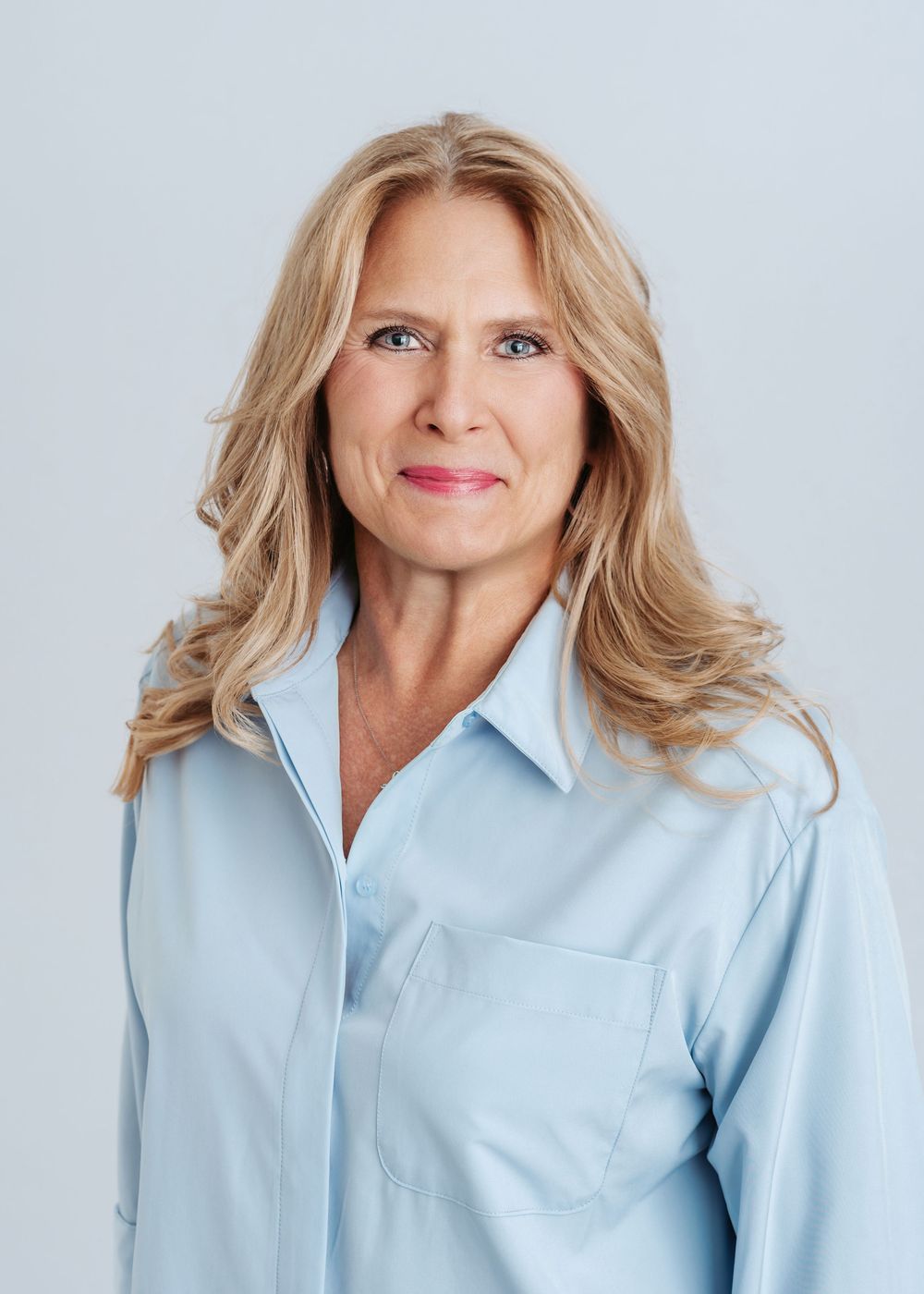 Professional headshot of a smiling blonde woman in a light blue button-up shirt against a gray background.