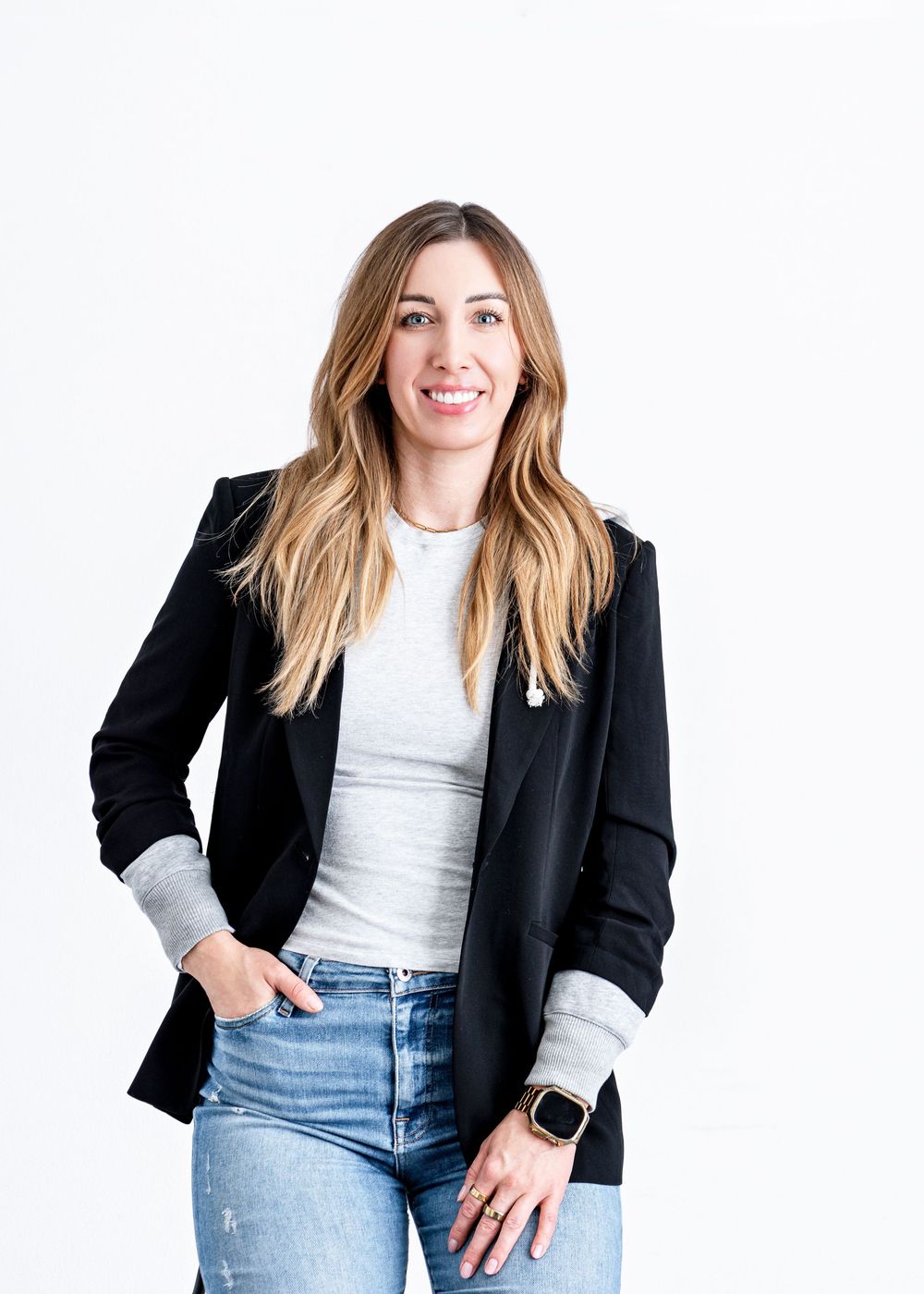 Woman in black blazer and jeans smiling confidently against white background, wearing a smartwatch.