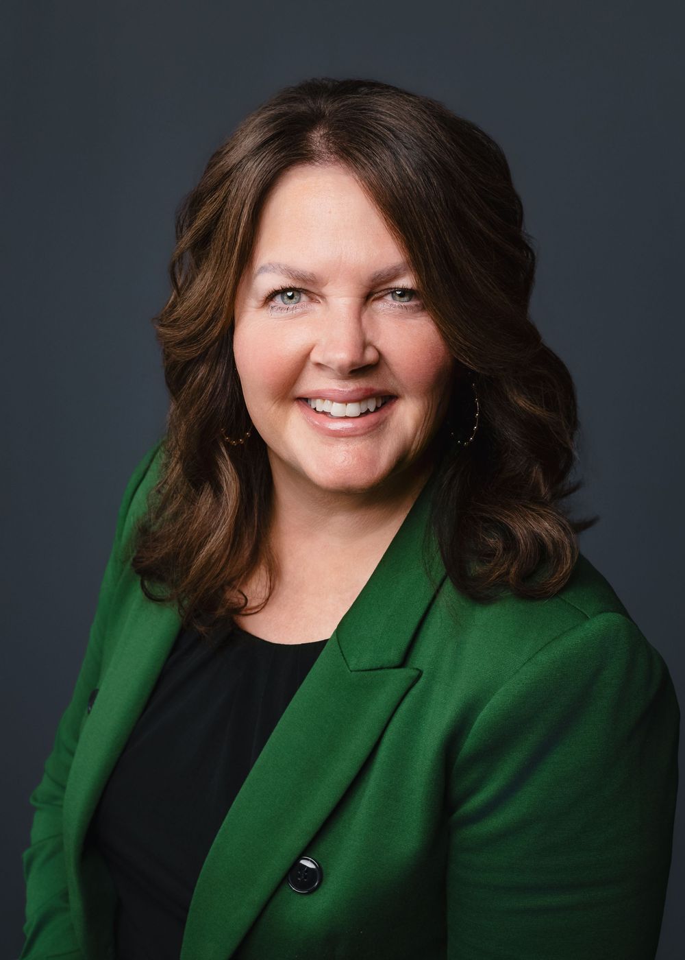 Professional headshot portrait of a person with a warm smile wearing a green blazer and black top against a dark background.