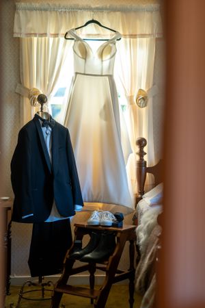Wedding attire including a white bridal gown and dark suit jacket displayed near a window with sheer curtains.