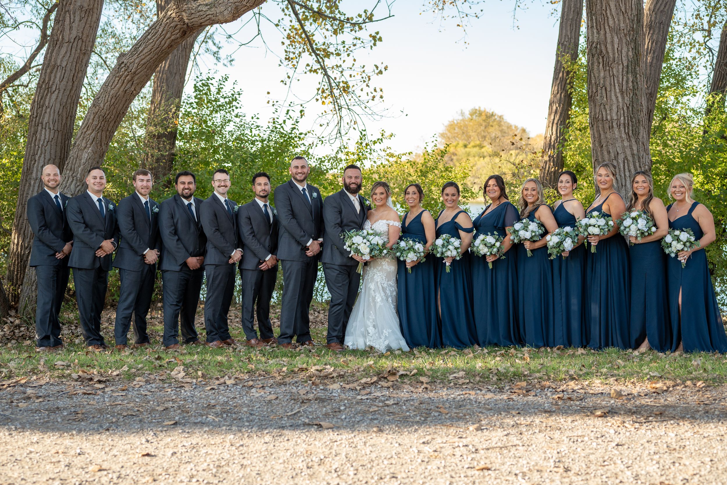 Wedding party in navy blue and gray attire poses together under tall trees at sunset.