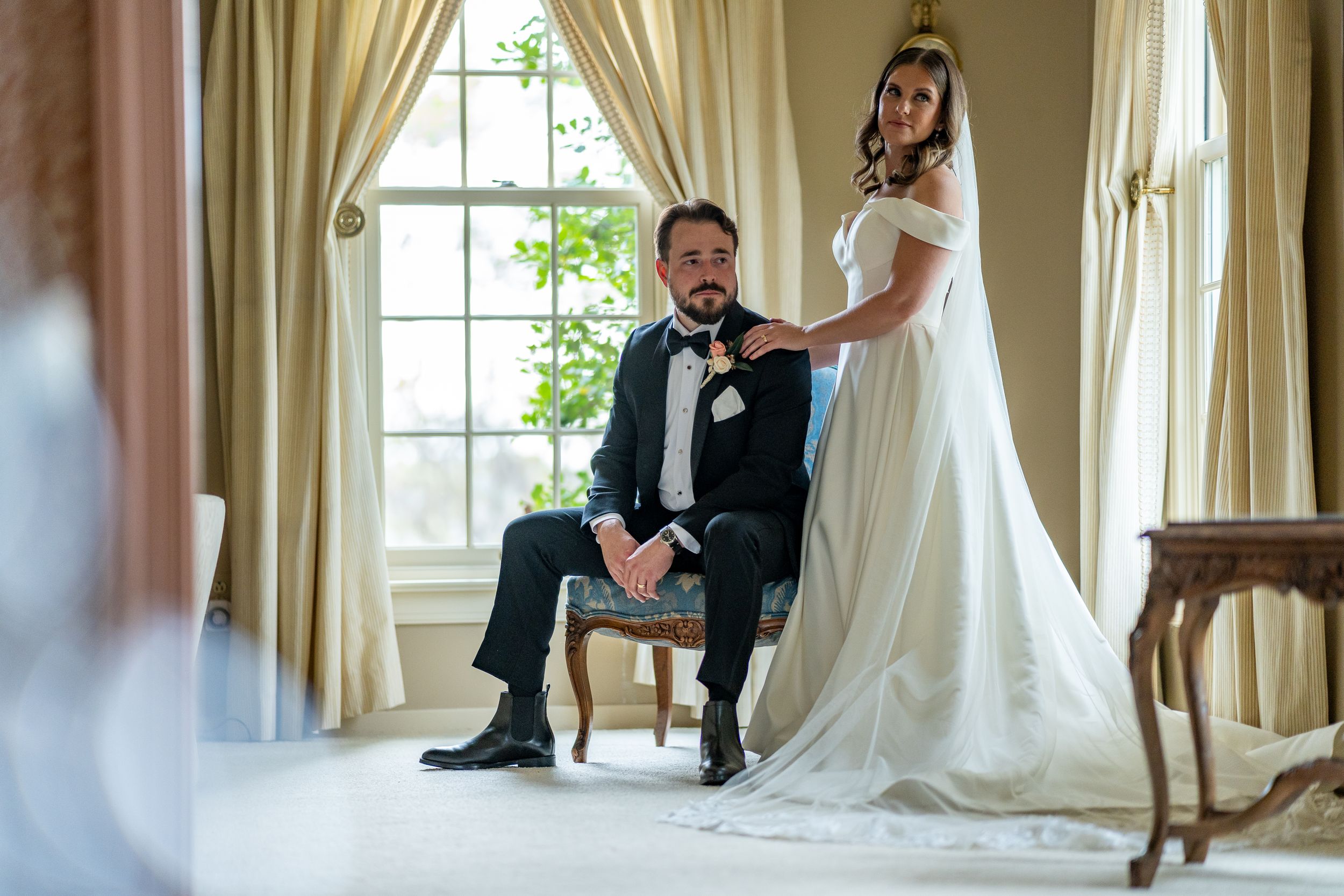 Elegant couple posing together in formal wedding attire by a window in classic interior setting.