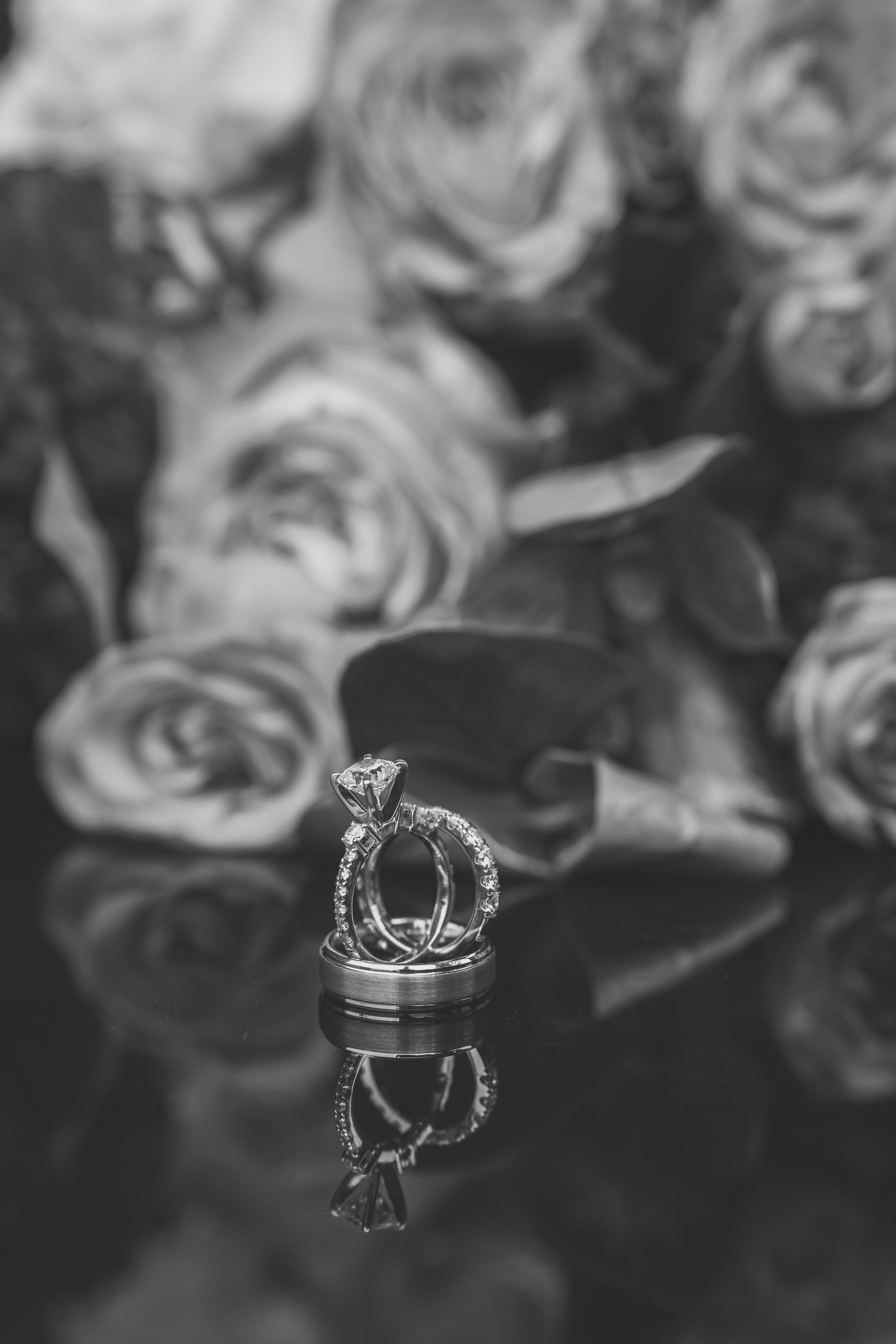 Wedding rings reflected on glossy surface with soft-focus roses in black and white background.