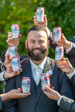 Wedding party member holds up multiple cans of beer while others join in with celebratory spirit.