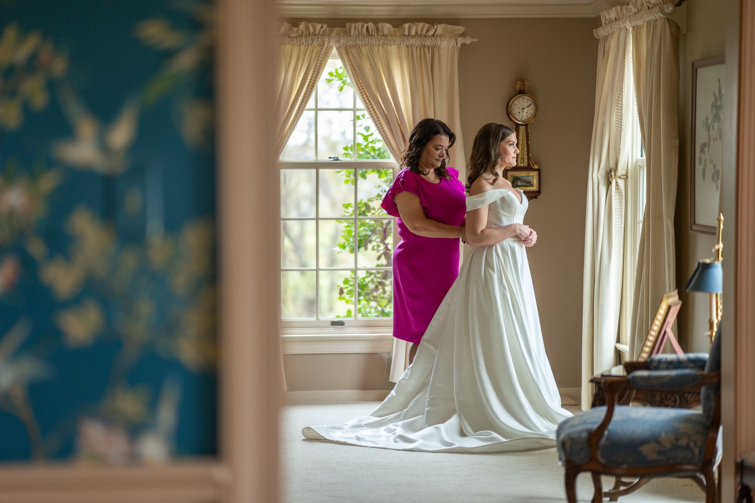 A wedding dress being adjusted in an elegant room with cream curtains and blue wall accents.