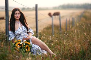 A beautiful woman sitting against a fence post with a basket of sunflowers and cows in the distance.