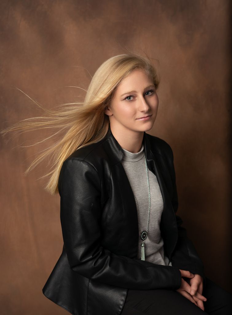 Professional studio portrait showing subject in black leather jacket and gray turtleneck against brown backdrop with hair in motion.