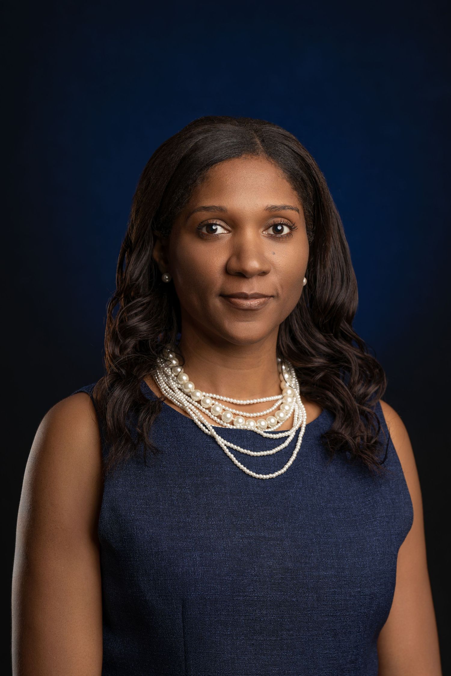 Professional headshot of person in navy dress with layered pearl necklaces against dark background.