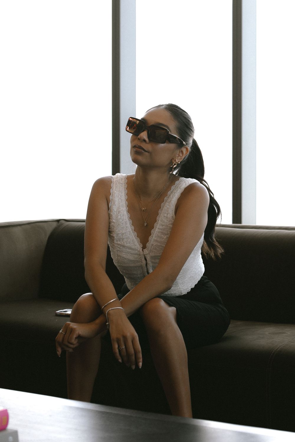 A person in sunglasses and white top sits on a dark couch in a contemplative pose near a window.