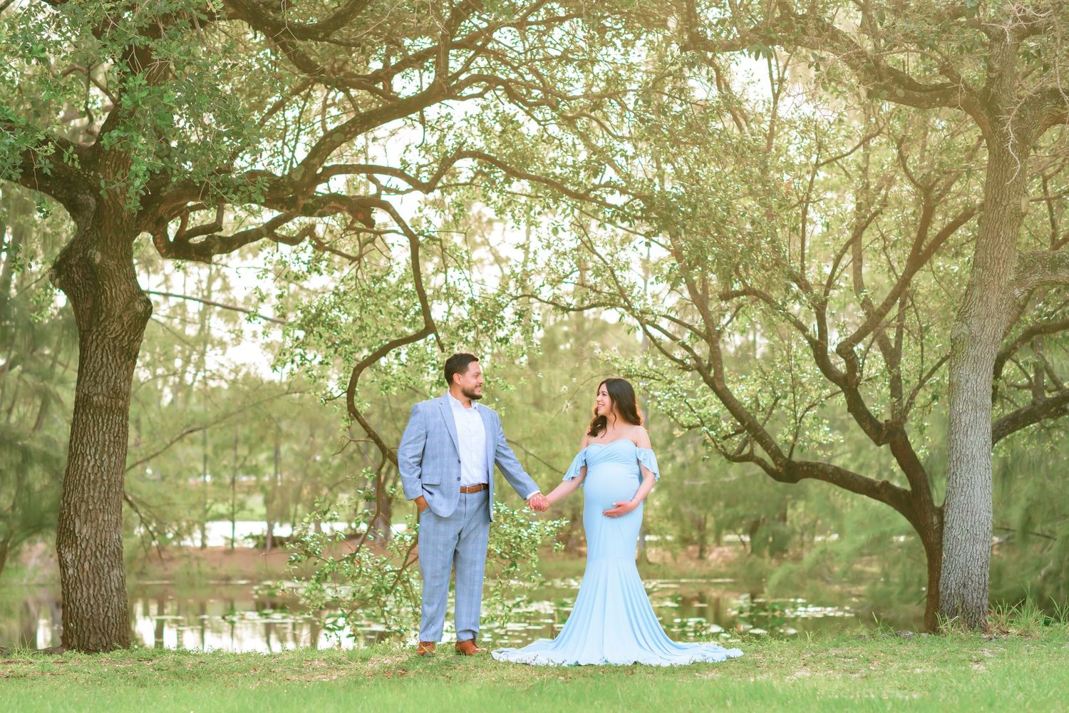 A couple in light blue outfits stands beneath moss-draped oak trees in a dreamy outdoor setting.