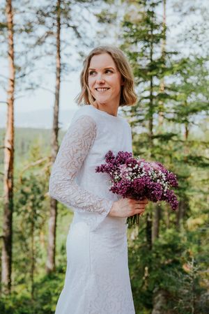Halvfigursbilde av bruden. Hun ser over skulderen og holder en bukett bestående av syriner og roser.