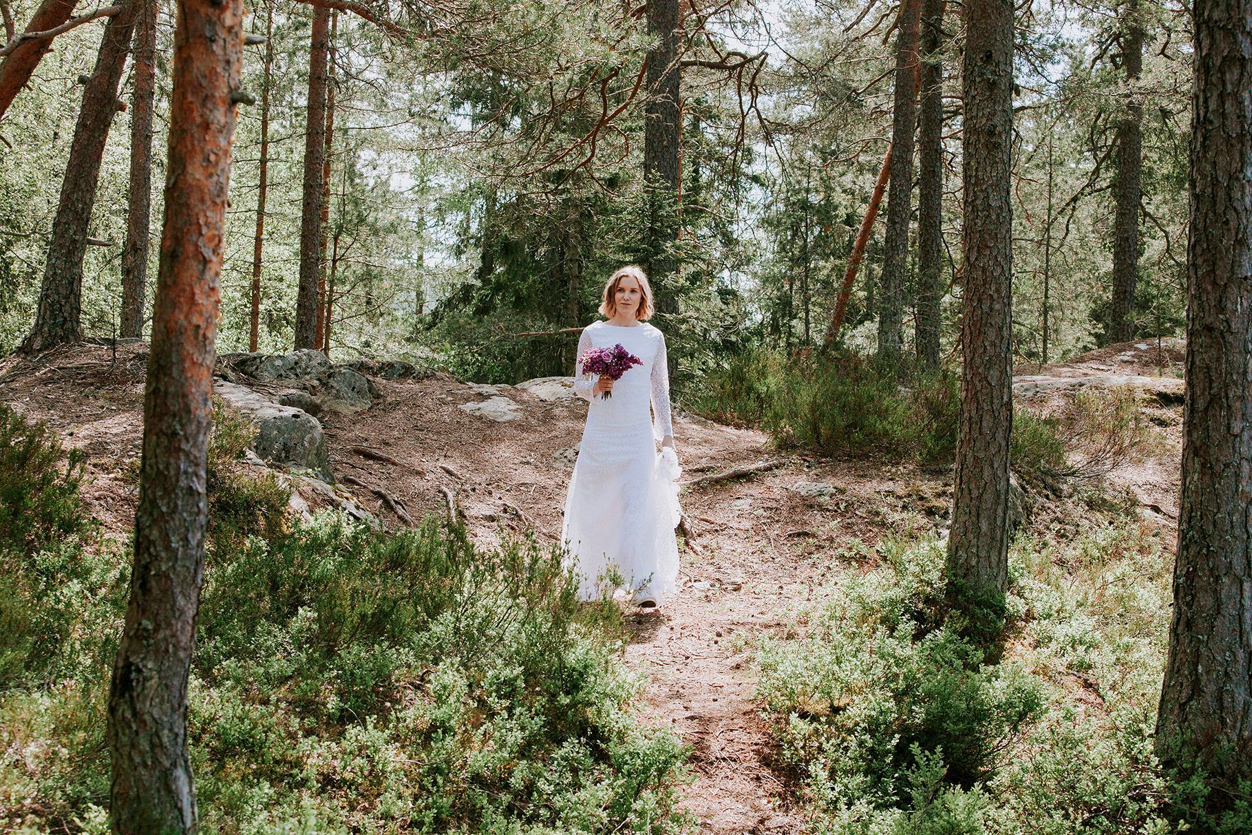 Bruden går gjennom skog med høye trær. Solen skinner litt igjennom og skaper et nydelig lys.
