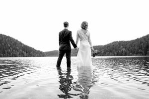 Brudepar med ryggen til kamera. Står i vannet med kjole og dress og speider utover mot utsikten av vann og skog.