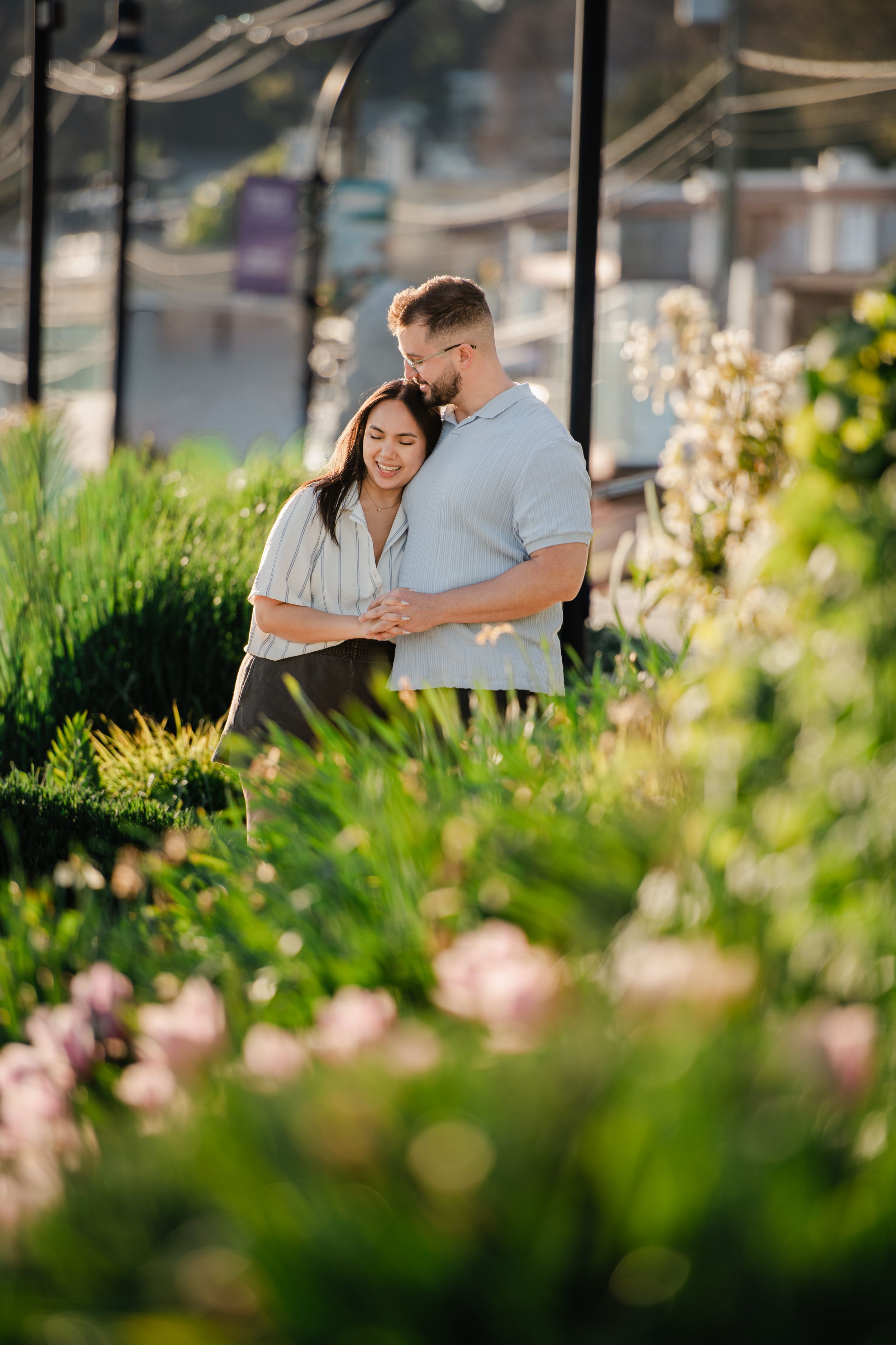 A couple embraces tenderly among lush green plants and pink flowers in a sunlit garden setting.