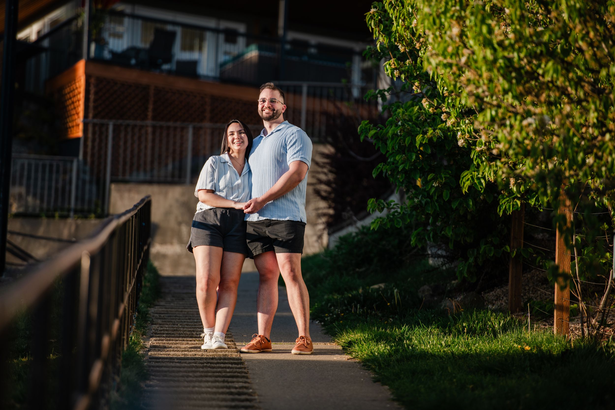 A smiling couple stands together on a sunlit pathway surrounded by lush greenery and rustic buildings.