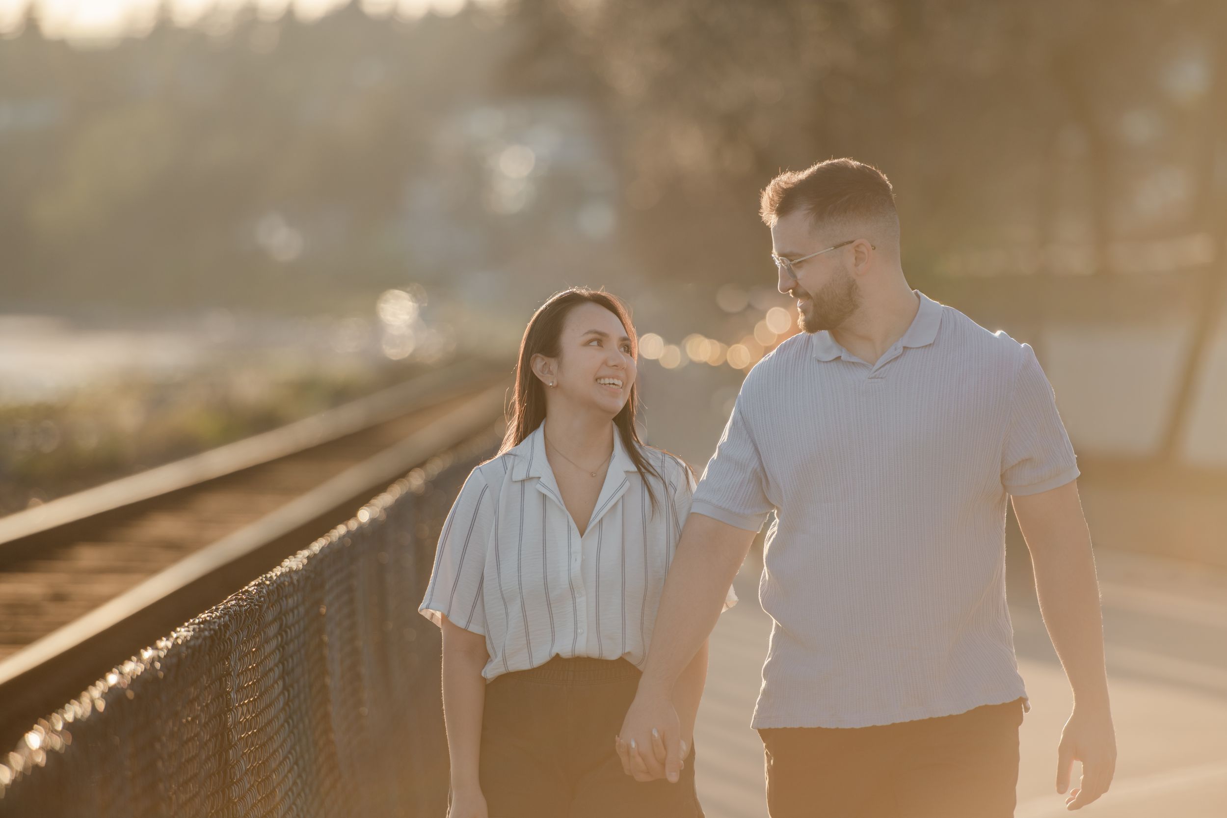 A couple walks hand in hand at golden hour near railroad tracks, smiling at each other in warm sunset light.