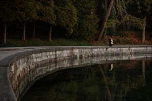 A lone figure jogs along a curved concrete wall next to a reflective pond in a dimly lit wooded area.