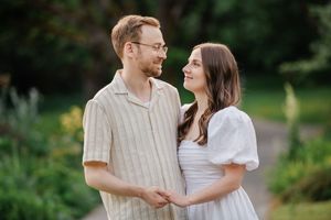 A couple wearing white outfits smile at each other while standing in a lush garden setting during golden hour.