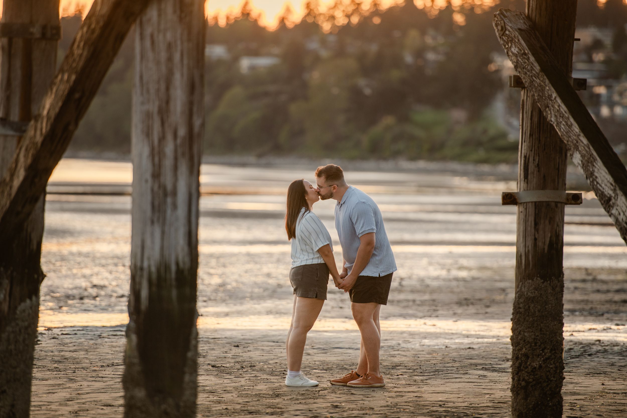 Couple sharing a romantic kiss on a sandy beach at golden sunset, framed by rustic wooden pier posts.