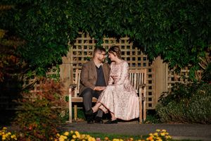 Couple sitting close on a wooden bench surrounded by lush green vines and colorful flowers in a garden.