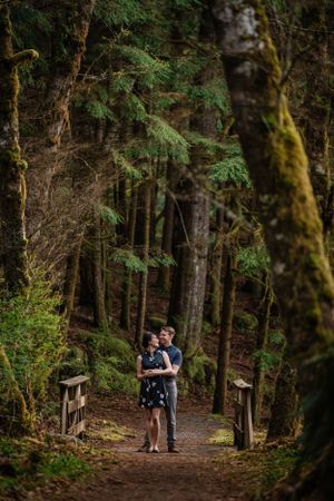 A couple embraces on a forest trail surrounded by tall evergreen trees and moss.