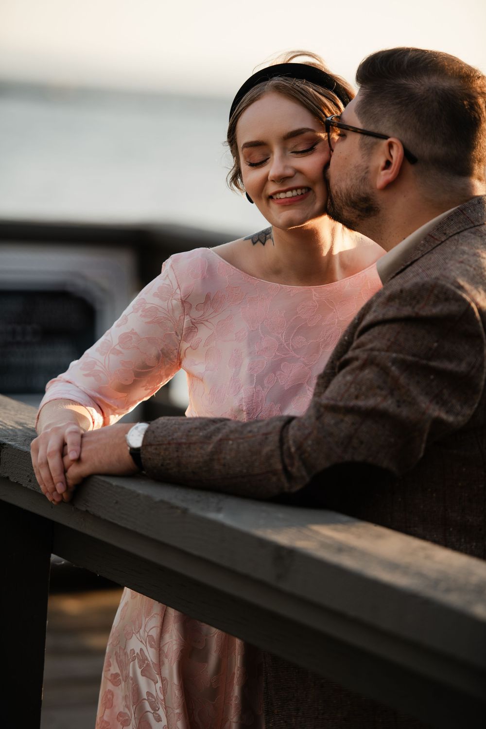 A couple shares a tender moment at sunset, man kissing woman's cheek as she smiles in a pink dress.