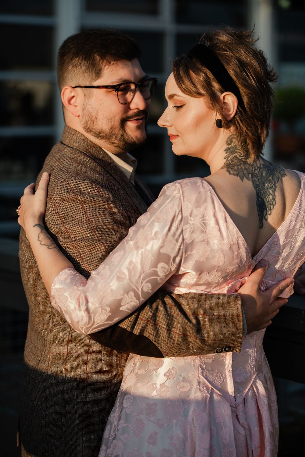A tattooed couple in elegant attire embraces tenderly outdoors, the woman in a pink floral dress and man in a tweed suit.