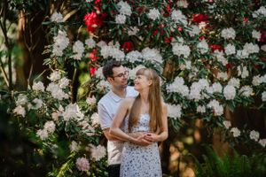 A couple shares a romantic moment in front of a wall of white and red flowers in a garden setting.