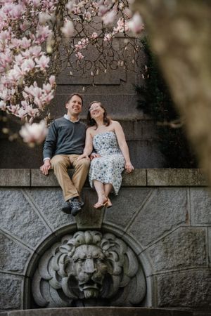 A couple sits on a stone wall with a decorative lion head carving beneath blooming pink magnolia branches in spring.