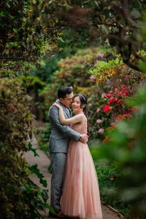 A romantic couple embraces on a garden path surrounded by vibrant red flowers and lush greenery.