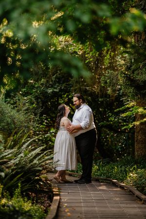 Couple embracing on a wooden boardwalk path surrounded by lush green garden foliage during an outdoor engagement session.