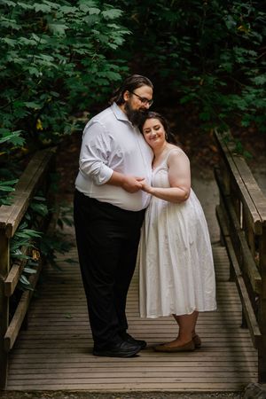 Couple embracing on a wooden bridge surrounded by lush green trees, woman in white dress leaning on man in white shirt.