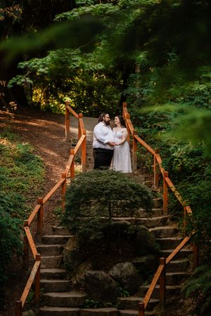 Couple in wedding attire embracing on wooden staircase surrounded by lush green forest trees.