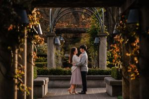 A romantic couple embraces under an ornate garden archway during golden hour with autumn foliage in the background.